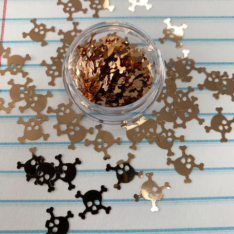 A clear container of Skull Shape Glitter for Halloween Nails Art Design by Dynamic Nail Supply sits atop a blue and white lined paper background, filled with metallic gold skull and crossbones confetti. Some glitter pieces are scattered around, catching the light to create a shiny effect.