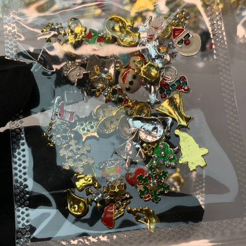 A gloved hand from Dynamic Nail Supply holds a transparent packet containing 50 pieces of small, Christmas-themed metallic charms. These charming accessories, ideal for winter nails art design, feature gold bells, snowmen, Santa faces, Christmas trees, and candy canes with festive red and green accents.
