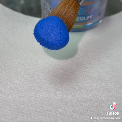 A paintbrush featuring a natural wooden handle and metallic ferrule holds a vibrant round puff of Dynamic Acrylic Dipping Powder S 007 - Solid Blue (Cobalt) by Dynamic Nail Supply. Below, a textured white paper towel is placed with a partially exposed container of nail art powder visible in the background. The TikTok app interface displays a username at the bottom right.