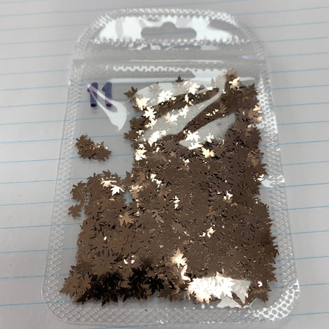 A clear plastic bag filled with tiny metallic gold star-shaped confetti pieces is placed on a lined notebook. Next to it, a bag of Dynamic Nail Supply's Fall Leaf Glitter features color-shifting maple leaves sequins for nail art design, with some spilling out and adding sparkle to the simple scene.