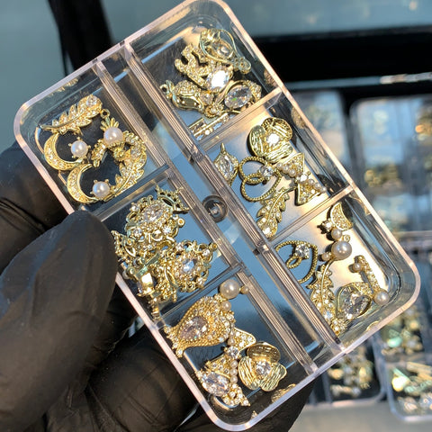A gloved hand holds a transparent rectangular box containing 30pcs of Dynamic Nail Supply's nail charms, each of the six compartments filled with intricate, random-shaped charms adorned with pearls and crystals. The designs feature bows and floral patterns. Another similar box can be seen in the blurred background.