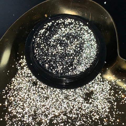 A mini container from Dynamic Nail Supply's Reflective Pixie collection releases shimmering silver glitter rhinestones onto a reflective black and gold surface. These glimmering particles capture the light, creating a dazzling super sparkle effect ideal for eye-catching nail art against the contrasting dark background.
