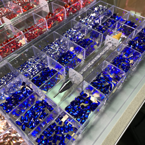 The Bling Bling set from Dynamic Nail Supply, featuring 3,000 pieces each of dazzling stones in rose, red, blue, and gold shades, is arranged neatly in a grid pattern. A rhinestone picker tool is placed diagonally across the containers. The stones catch the light beautifully, producing a vibrant and colorful spectacle.