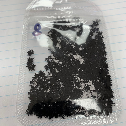 A clear plastic bag featuring color-shifting maple leaves sequin from Dynamic Nail Supply, complete with a small purple infinity symbol, rests on a lined notebook page. The glitters inside, reminiscent of black star-shaped confetti and fall leaf glitter, are scattered across the sealed bag, showcasing their shapes against the paper background.