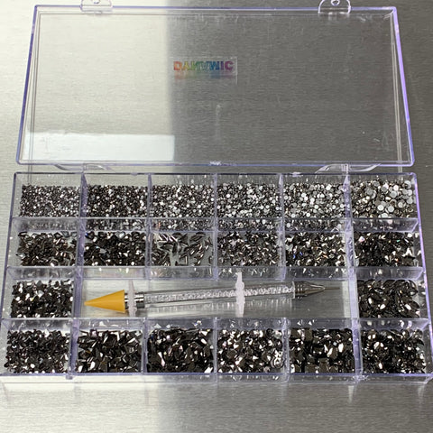 The Gun Metal Color Rhinestones set from Dynamic Nail Supply features a clear plastic organizer box with a transparent lid, housing a variety of metallic nail art gems, including 14 big shapes and 6 round shapes. The box is divided into 20 compartments, and it comes with a nail art picker tool that has a yellow cone tip and a silver pen-like body positioned across the middle.