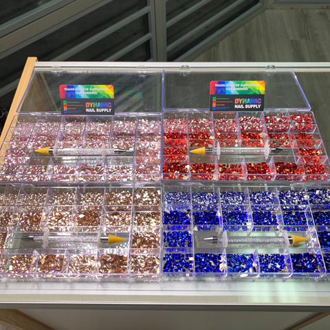 A well-lit room with large windows features a glass display case showcasing nail art supplies. Inside are four trays filled with the Bling Bling set from Dynamic Nail Supply, featuring colorful rhinestones in pink, red, and blue. Each tray is organized into small compartments with a neatly placed rhinestone picker on top.