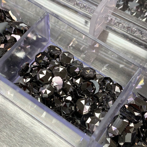 A clear plastic container from Dynamic Nail Supply showcases an assortment of shiny, faceted rhinestones in a Gun Metal color. These stones create sparkling reflections under the light. The background hints at more rhinestones stored in additional compartments, featuring 14 big shapes and 6 round shapes within the visible batch of this 20-cell Gun Metal color Rhinestones set.
