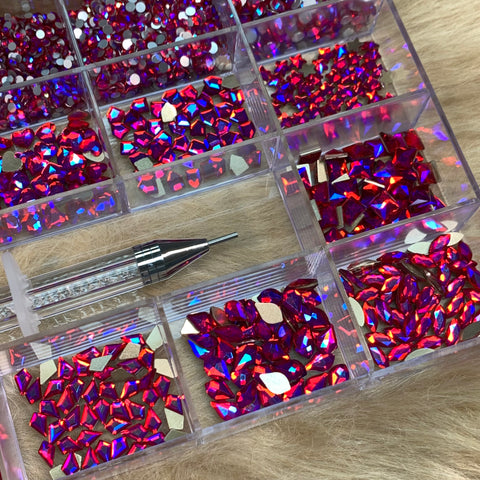 A close-up of a [iridescent magenta] Rhinestones set from Dynamic Nail Supply, housed in a clear, compartmentalized 20-cell box featuring 14 large shapes and 6 round shapes, showcases vibrant red and iridescent magenta gems against a soft, beige fur backdrop. An essential for nail enthusiasts, the pointed metal-tip picker tool with its transparent handle is poised for precision artistry.