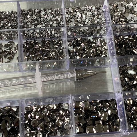 A transparent organizer from Dynamic Nail Supply contains 20 compartments filled with gleaming gun metal rhinestones, featuring 14 large shapes and 6 round shapes. Resting atop the box is a tool with a clear cylindrical handle and a pointed metal tip, ideal for applying these faceted stones that beautifully catch and reflect light.