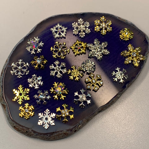 Displayed on a glossy, dark blue agate slice is a set of 24 intricately designed metallic snowflake charms from Dynamic Nail Supply, perfect for enhancing diverse nail shapes. Featuring silver and gold hues, these charms vary in size and design, some adorned with embedded gemstones for an added touch of sparkle.