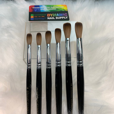 A set of six Dynamic Kolinsky Hair Brushes, designed with black handles and featuring 100% pure crimped Kolinsky hair for acrylic nail application, is displayed on a fluffy white surface. Behind the brushes, a colorful business card for Dynamic Nail Supply can be partially seen, containing contact information and a thank you message.