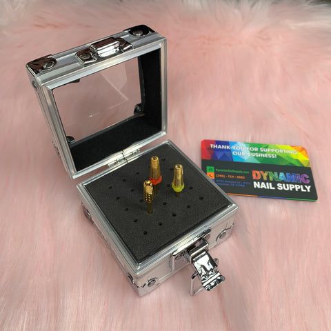 A small container box for drill bits from Dynamic Nail Supply, featuring a silver aluminum finish with an open lid, reveals the drill bit holder with bits neatly inserted into a foam pad. The interior is lined with black material. Two colorful business cards from Dynamic Nail Supply are placed beside the case on a fluffy pink surface.