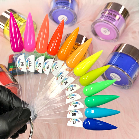 A fan of the Dynamic Nail Supply's (Part 2) 2024 Summer Acrylic Collection features a vibrant array of glossy colors, spanning from pink to blue. Made with non-toxic ingredients, these long-lasting nails are marked with specific color codes. In the background, blurred jars can be seen as a black-gloved hand gracefully holds the fan.