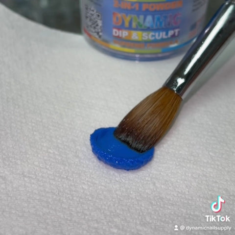 A makeup brush sweeps vivid cobalt blue powder onto a textured white surface, achieving the ideal look for dipping nails. In the background, a container of Dynamic Acrylic Dipping Powder S 007 - Solid Blue (Cobalt) can be seen. The TikTok and Dynamic Nail Supply logos appear at the bottom right corner.