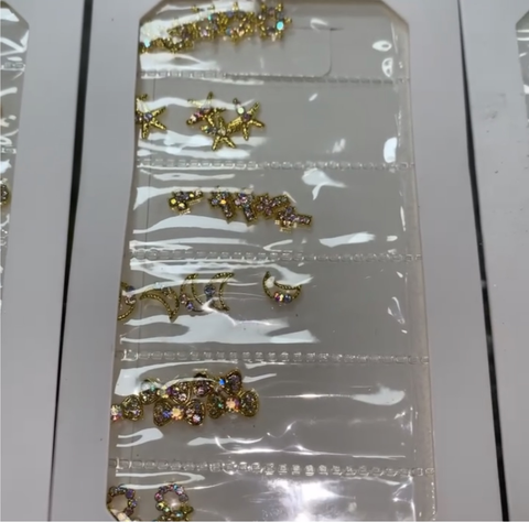 Displayed is a sheet featuring Pre-made Nail Charms from Dynamic Nail Supply, showcasing gold designs like stars, moons, flowers, and bows. Some pieces are adorned with colorful rhinestones. These elegant designer nail elements are arranged in rows on a clear plastic sheet within a white frame.