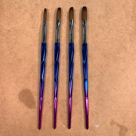 Four [Crimped] Dynamic Kolinsky Brushes by Dynamic Nail Supply feature metallic handles that transition from blue to pink, ideal for acrylic nail applications. The rounded, brown bristles are crafted from 100% pure Kolinsky hair. These brushes are neatly lined up parallel on a textured brown surface.