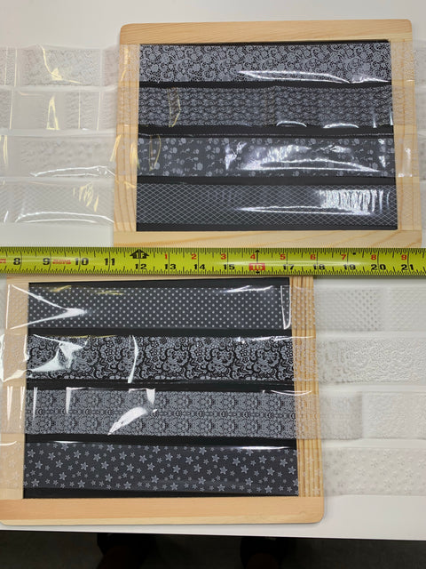 Two wooden frames wrapped in clear plastic showcasing the New Designer Foils, featuring black fabric with a variety of white patterns including polka dots, stripes, floral, and star motifs. A yellow measuring tape runs horizontally above the style #001 frames. Part of a textured white material is visible next to the Designer Foils from Dynamic Nail Supply.