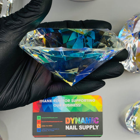 A gloved hand holds a large, multifaceted K9 Crystal resembling a diamond with blue reflections. Below, a colorful business card displays the text "Thank you for supporting our business! Dynamic Nail Supply" along with contact information, surrounded by exquisite 10cm (4") Display Diamond - K9 Crystals as delicate table decorations.