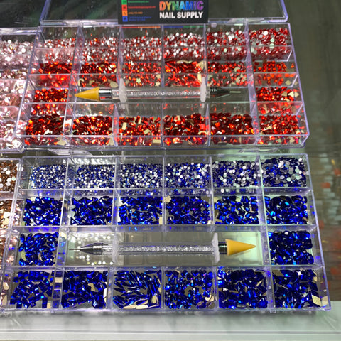 An organized display of nail art rhinestones in clear plastic containers showcases the Rose, Red, Blue, and Gold shades from the Dynamic Nail Supply "Bling Bling set," each containing 3000 pieces. A dual-ended rhinestone picker with a crystal-filled handle by Dynamic Nail Supply is prominently placed in the center, beneath signs that read "Bling Bling sets.
