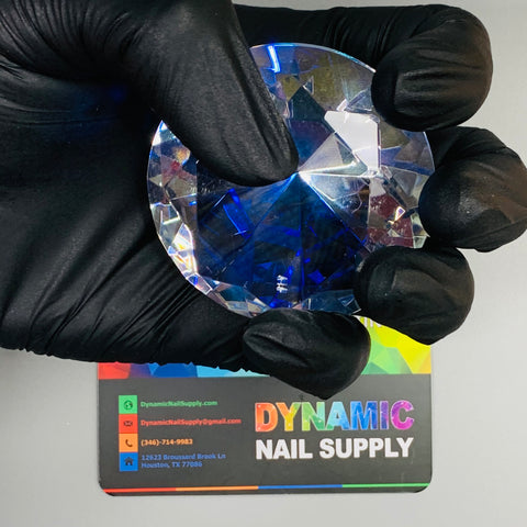 A gloved hand holds a 6cm (2.3") Display Diamond made from K9 crystal, featuring a blue tint. Below it, a colorful business card for Dynamic Nail Supply displays contact information such as an email address, phone number, and Houston, TX address against a vibrant rainbow gradient backdrop.