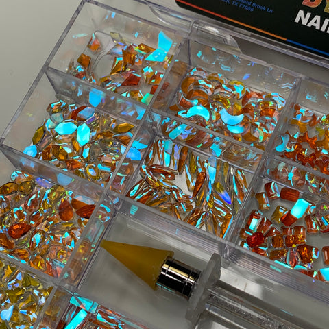 A divided container from Dynamic Nail Supply displays a selection of 1,000 iridescent rhinestones in captivating hues like blue aurora and clear crystal. Perfect for dazzling nail art designs, each stone features a reflective, holographic effect. Alongside these rhinestones is a yellow-tipped, angled crystal picker tool for precise placement against the packaging backdrop.