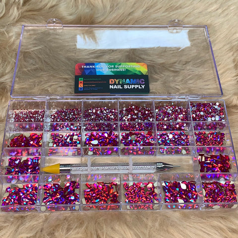 A plastic organizer from Dynamic Nail Supply, designed with transparency and multiple compartments, beautifully displays the [iridescent magenta] rhinestones set. This set includes 20 cells filled with gems in 14 big shapes and 6 round shapes. A dual-ended nail tool is elegantly positioned across the center of the organizer. On its lid lies a card from Dynamic Nail Supply, all set against a plush beige background.