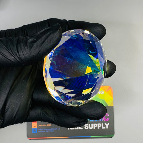 A gloved hand holds the 6cm (2.3") Display Diamond from Dynamic Nail Supply, a large, multifaceted K9 crystal that exhibits shades of blue and yellow. This captivating piece is showcased above a partially obscured colorful business card, with a plain light gray background enhancing the display of this exquisite decoration.