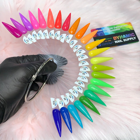 A gloved hand presents a set of vibrant, pointed nail polish samples from the 2024 Summer Acrylic Collection by Dynamic Nail Supply, elegantly arranged on a metal ring against a plush pink surface. The color spectrum ranges from pinks and reds to greens and blues. In the background, a card partially reveals the Dynamic Nail Supply branding, emphasizing their non-toxic acrylic powders designed for long-lasting nails.