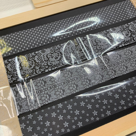 A zoomed-in view highlights a display featuring the New Designer Foils, style #001, presented in a wooden frame. These translucent black foils from Dynamic Nail Supply boast captivating patterns of polka dots, floral lace, intricate filigree, and stars. The glossy surface captures and reflects light beautifully, revealing elegant reflections and highlights.