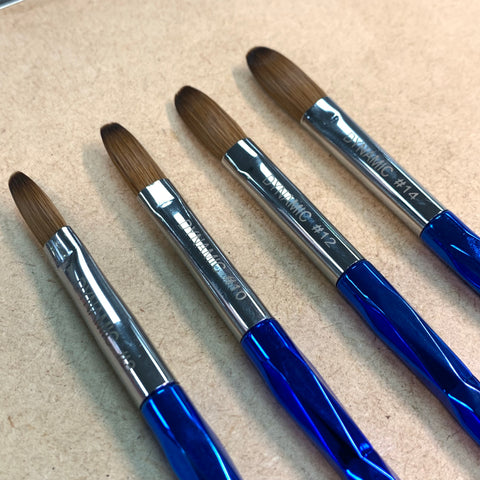 Four Dynamic Nail Supply [Crimped] Dynamic Kolinsky Brushes, featuring blue handles in sizes 8, 10, 12, and 14, are arranged on a beige wooden surface. These brushes are perfect for acrylic applications with shiny metal ferrules and uniform brown bristles. The sleek and pointed tips make them ideal for detailed work.