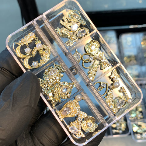 A gloved hand holds a plastic case from Dynamic Nail Supply containing their 30pcs (Random Shapes) Nail Charms designed for nail art and decoration. These gold-colored charms feature rhinestones, pearls, and intricate designs, all neatly arranged in compartments. The background shows similar cases filled with a variety of decorative charms.