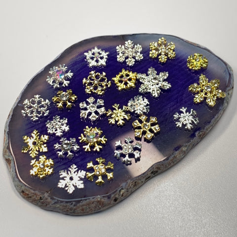 A flat, polished stone slab from Dynamic Nail Supply elegantly showcases the 24pcs Snowflake Charms for Winter and Christmas nails art design. The gold and silver snowflakes, intricately detailed to mimic elegant nail shapes, are neatly arranged across the surface, while the slab maintains a rough, natural edge.