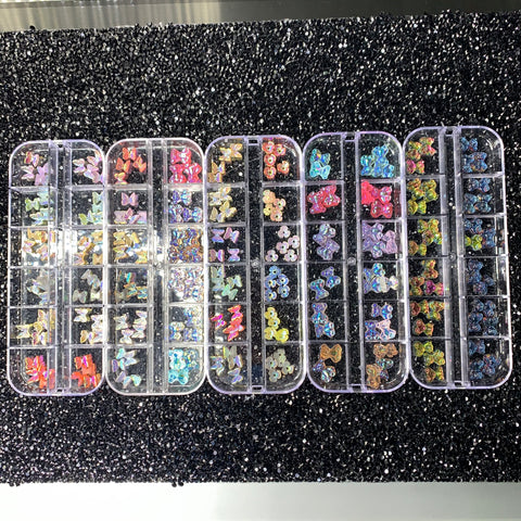 Five transparent containers from Dynamic Nail Supply, featuring an assortment of colorful gummy butterflies along with flower charms and butterfly-shaped nail art decorations, are artfully displayed on a black, textured surface. Each container is divided into small compartments that showcase a variety of colors and designs, including metallic and holographic patterns.