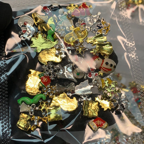 A clear plastic bag from Dynamic Nail Supply, featuring their "50 pcs Christmas Charms for Winter Nails Art Design," includes an assortment of small, colorful charms such as snowmen, gold reindeer, silver stars, and green Christmas trees. These intricately detailed charms are perfect for nail art design and sparkle against the light in a gloved hand.
