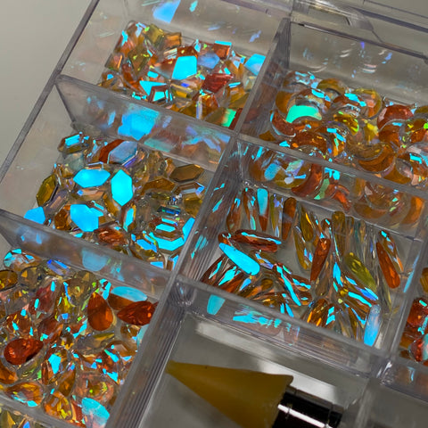 Dynamic Nail Supply's 1000 pcs Iridescent Blue Aurora Clear Crystal Rhinestones set features a clear plastic organizer filled with vibrant, iridescent rhinestones and crystals in an array of shapes and sizes, ideal for nail art enthusiasts. The stones captivate with their stunning orange and blue hues, while a tool with a yellow tip is conveniently positioned at the bottom.