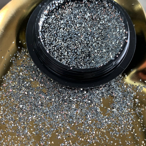 A close-up of an open container reveals Reflective Pixie (mini sparkle rhinestone) by Dynamic Nail Supply, silver holographic glitter spilling onto a shiny gold surface. These reflective pixie pieces are small yet dazzle with a super sparkle, perfect for creating enchanting nail art with their sparkly, multicolored effect.