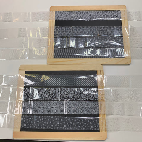 Two wooden-framed slate boards rest on a white surface, each adorned with intricate black and white patterned strips reminiscent of Dynamic Nail Supply's New Designer Foils (style #001). Transparent plastic sheets partially cover the boards, lending a glossy effect that beautifully contrasts with the matte textures underneath.