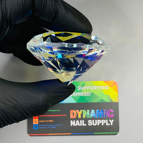 A gloved hand holds a 6cm (2.3") Display Diamond - K9 Crystal Diamond from Dynamic Nail Supply over a colorful business card. The card, featuring a rainbow gradient with text in white and black, includes contact information. This stunning display diamond rests against a plain, light surface.