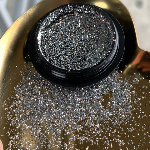 A petite container in black from Dynamic Nail Supply rests on a golden surface, overflowing with their Reflective Pixie mini sparkle rhinestone. Some of the silver glitter has spilled, illuminating the scene akin to the enchanting pixie dust. The gold backdrop enhances the shimmer of the glitter, making it ideal for captivating nail art lovers.
