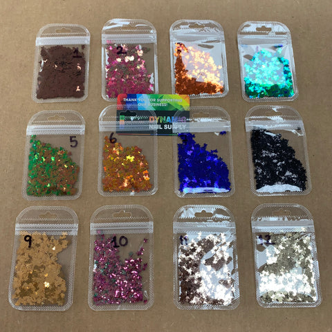 Arranged in four rows on a brown surface are twelve small plastic bags, each filled with vibrant Fall leaf glitter and Maple leaves sequins designed for nail art. Every bag showcases a unique color that shifts under different lighting and is labeled numerically from 1 to 12. At the center proudly sits a business card for Dynamic Nail Supply.