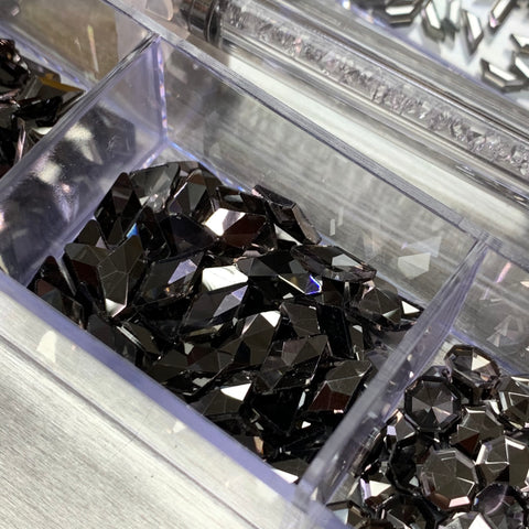 A close-up reveals a clear box filled with multiple rhinestones from the Dynamic Nail Supply Gun Metal color Rhinestones set. This 20-cell container showcases diamond-shaped gems with angular facets that brilliantly reflect the light. Nestled in transparent compartments on a metallic surface, additional stones from this set sparkle in adjacent sections.