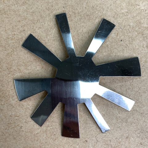 A shiny metallic object with eight flat, reflective blades arranged in a radial pattern, resembling a star or sunburst, lies on a plain, textured beige surface. Known as the French Nails Cutter by Dynamic Nail Supply, its metal surface catches the light, creating bright reflections on each blade.