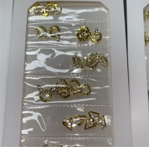 A close-up display features a stunning collection of Pre-made Nail Charms from Dynamic Nail Supply, highlighting intricate designs and embedded sparkling gems. Neatly arranged in transparent pockets, the assortment includes hoops, triangles, and circular patterns that reflect light beautifully against a white background.