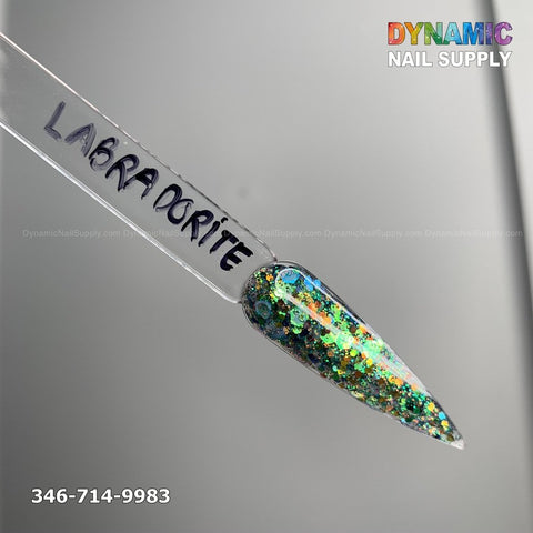 A nail sample named Labradorite #329 - Acrylic Glitter Powder, featuring a pointed tip, exhibits chip-resistant and iridescent colors, predominantly green and blue, enhanced with silver glitter. The simple background brings attention to the Dynamic Nail Supply logo in vibrant text at the top right and a phone number at the bottom left.