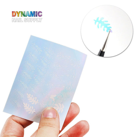 A hand holds a translucent sheet of nail art stencils with intricate leaf patterns. In the top right inset, a small paintbrush is applying turquoise polish through a stencil, producing an eye-catching leaf design. The text "Dynamic Nail Supply" appears in colorful lettering at the top left, highlighting their Hot Design 16pcs/set - Holographic Leaves Sticker for all your nail art decoration needs.