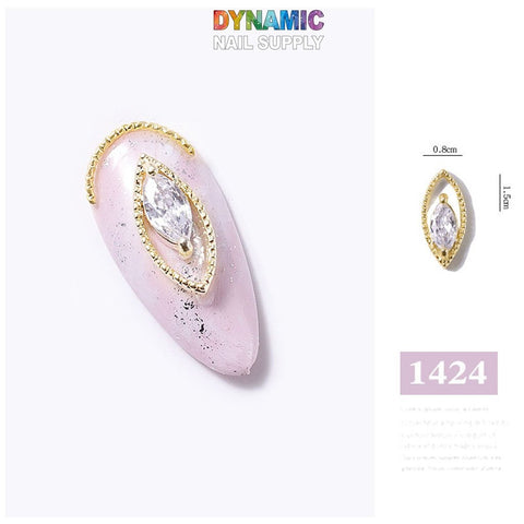 This image showcases a luxurious nail art design from Dynamic Nail Supply, specifically their "Luxurious Jewelry for Nail Art design - part 4." The design features a light pink base adorned with black speckles. At the center is a gold, leaf-shaped charm with a marquise-cut crystal, topped by a gold arc accent. The dimensions are 0.8 cm in width and 1.5 cm in height, with the number "1424" visible as part of the display.