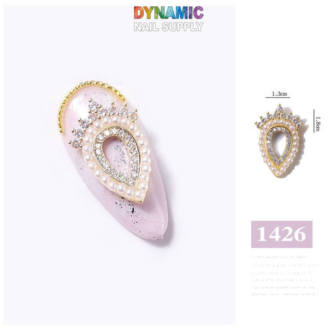 In this exquisite nail art design, a close-up showcases a luxurious teardrop-shaped charm from the Luxurious Jewelry for Nail Art Design - Part 4 collection on a light pink nail accented with black speckles. The gold charm is resplendent with pearl and diamond-like embellishments. Above, the Dynamic Nail Supply logo is prominently featured, with "1426" elegantly presented in purple text on the right.