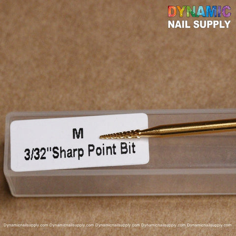 Resting on its transparent plastic case, the Medium Sharp Point Bit - Dynamic Drill Bit, ideal for sealing the cuticle area and featuring a gold color, displays its label M 3/32 Sharp Point Bit. Against a beige background, the colorful text of Dynamic Nail Supply is prominently featured at the top right corner.