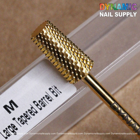 A close-up shows the gold-colored M Tapered Barrel Bit, a Dynamic Drill Bit, resting on a beige surface. The background features a transparent plastic case with "Dynamic Drill Bit" clearly labeled. In the top-right corner, the logo of Dynamic Nail Supply is prominently displayed in vibrant text.