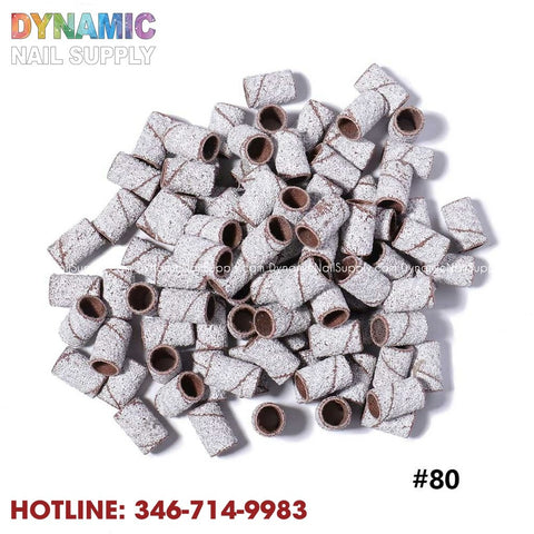 A collection of cylindrical sanding bands is shown against a white background, showcasing their textured, grey surface ideal for nail filing. The name "Dynamic Nail Supply" and the hotline number 346-714-9983 #80 are printed in vibrant text, making these Machine Nail Drill Bits Zebra Sanding Bands a seamless addition to your manicure toolset.